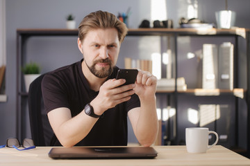 Happy bearded man in casual clothing using smartphone while sitting at table. Handsome male reading working messages on mobile at home.
