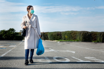 Ragazza con mascherina verde, cappotto chiaro , borsa nera  e una busta blu  cammina in mezzo alla...