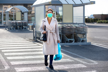 Donna con mascherina verde, cappotto chiaro , borsa nera  e una busta blu  cammina sulle strisce...