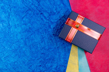 Closeup of a blue gift box with heart and red ribbons over color papers in background. Wives and...