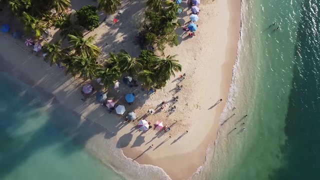 Top down shot from ascending drone of U shaped Venezuelan beach coastline. Parasols, palm trees and a few people cast evening shadows on the sand and sapphire green waters.