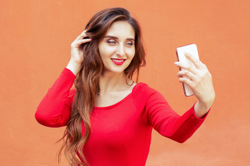 Beautiful young woman is taking selfie by smartphone on orange background.