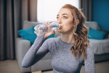 Portrait of charming young lady with blond hair knot drinking water during morning workout. Sporty girl in activewear refreshing after regular exercises at home.