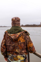 Man in camouflage trousers and jacket with hood catches fish on river in a cloudy day