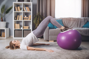 Full length portrait of active female in white t-shirt and grey leggings holding legs on fitness ball and doing exercises. Concept of healthy lifestyle and body activity
