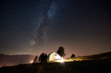 Tourist camping at summer night. Illuminated tent and campfire under magical night sky full of stars and Milky way. On the background beautiful starry sky, mountains and luminous town