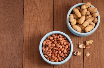 Two blue ceramic bowls of nuts. In one bowl there are peanuts in the shell, and in the other there are peeled nuts. Next to the bowls are peeled peanut kernels on a brown wooden table. Top view.