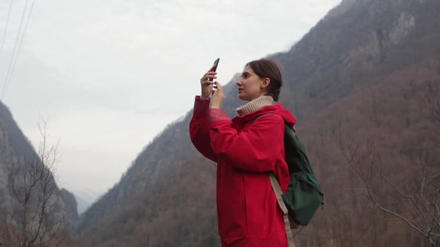 A girl in a red raincoat walks and takes photos of the mountains on her phone
