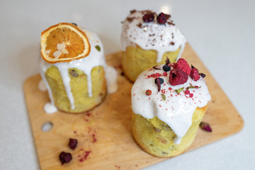 Various Easter cakes with white icing, decorated with berries and orange stand on a wooden board.