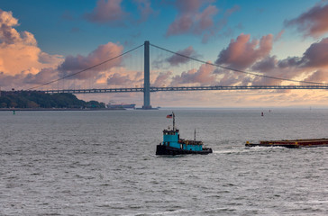 The Verrazano Bridge under grey skies on a foggy day in New York City with a blue tugboat pulling a barge