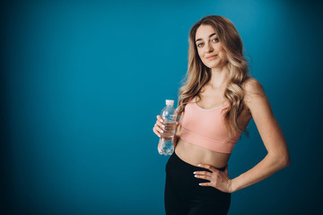 Portrait of charming woman in sport clothing posing in studio with bottle of water. Fitness girl with blond hair isolated over blue background.
