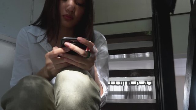 Depressed young beautiful Asian woman sitting alone on the stairs and using smartphone with internet. Stressed loneliness teenage girl looking at smartphone with negative emotion and closing her eyes.