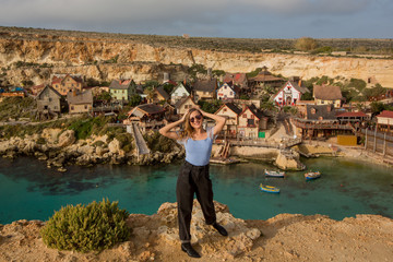 Fototapeta na wymiar Blonde Traveler girl on the background of Village. A panoramic view of the Village at Anchor Bay with traditional boats, beautiful colorful clouds and sky