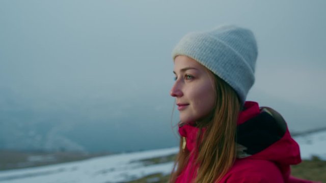 Young Smiling Female Adventurer in Red Jacket Standing on Top of Mountain Looking into Distance. Happy-Looking Girl Hiking, Exploring Nature and Enjoying Beautiful Views. Slow Motion Shot