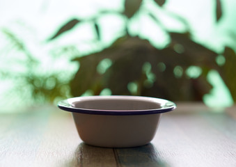 
White metal bowl on top of a wooden table with a background with plants.