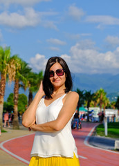 Positive woman walking and enjoys sea vacation and sunshine. Cheerful, smiling, lucky lady in white blouse, yellow trousers and sunglasses, outdoors near tropical palms. Sunny summer day.