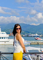 Fototapeta na wymiar Positive woman walking at the harbor on vacation. Mountains on background. Cheerful, smiling, lucky lady in white blouse, yellow trousers and sunglasses, outdoors at sea port. Sunny summer day.