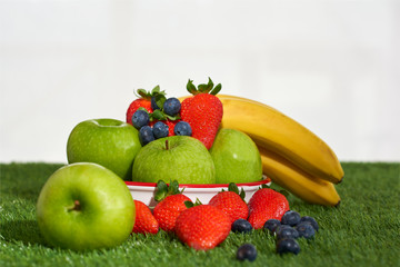Bodegon de fruta sonbre un cesped de color verde y un fondo blanco. En la imagen hay fresas, arandanos, manzanas, platanos.