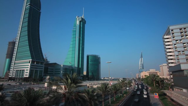 Tilting up moving timelapse of the King Faisal Highway in Manama with cars speeding in the foreground and the iconic Bahrain Financial Harbour twin towers in view with palm trees in the median.