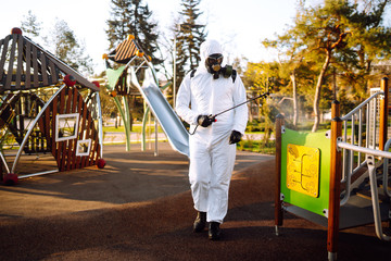 Man wearing protective suit disinfecting the playground in the sun with spray chemicals to preventing the spread of coronavirus, pandemic in quarantine city. Covid -19. Cleaning concept.