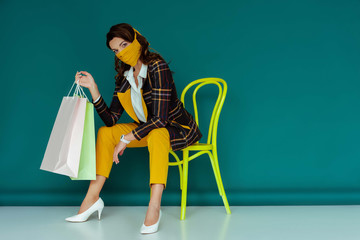 young woman in yellow mask and plaid blazer posing while sitting on chair with shopping bags on blue