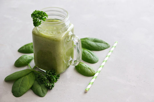 A glass mug, a glass, a glass with a zeon fresh smoothie of banana, apple, celery and spinach with leaves scattered next to it and a reusable straw on a gray concrete background. Healthy snack