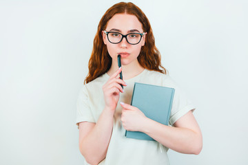 redheaded serious woman in eyeglasses holding pen in one hand near face and planner in another standing on isolated white backgroung, learning and teaching concept