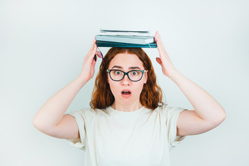 redheaded confused woman in eyeglasses with books on her head looks confused standing on isolated...