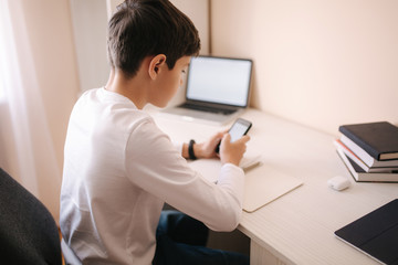 Fototapeta premium Schoolboy study at desk in his room. Boy use laptop and smartphone to writing in notebook. Books and tablet on the table. Study home during qurantine