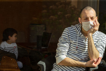 Man wearing face mask sitting by the window and his child during their self-isolation at home as infections and coronavirus disease are spreading. Suffering ill people at home to prevent an epidemic