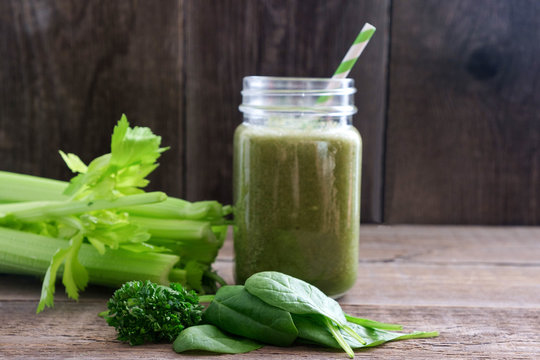 A glass with a green smoothie and a bamboo cocktail straw is standing on a wooden board. Nearby are bunches of celery and spinach and parsley leaves. Useful snack. Vegan, vegetarian drink. Copy space