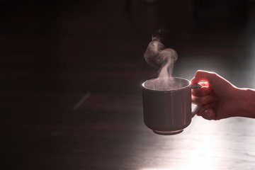 Silhouette of a female hand with a mug of hot drink - coffee or tea with steam on a black background with an empty place for text - horizontal shot