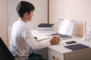 Schoolboy study at desk in his room. Boy use laptop for lessons. Drink tea. Books and tablet on the table. Study home during qurantine