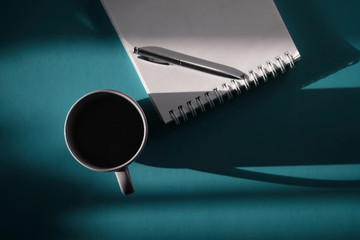 A hot drink in a mug pot on a cyan background on the floor - coffee or tea next to a notebook and pen. Flatlay workspace with empty space for text