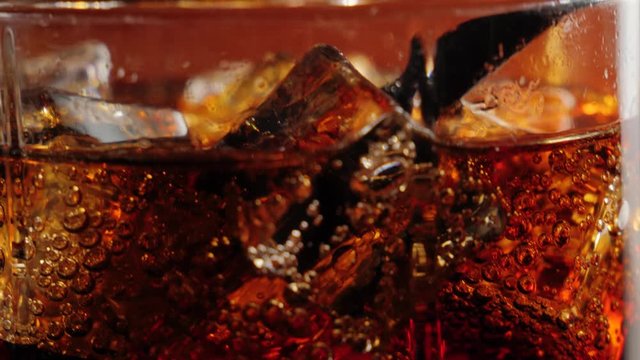 Camera moves up from ice cubes with bubbles in a glass of cola with black straw and brown highlights against black background. Concept of drinking coke or soda, relax and having fun. Close up. 4K