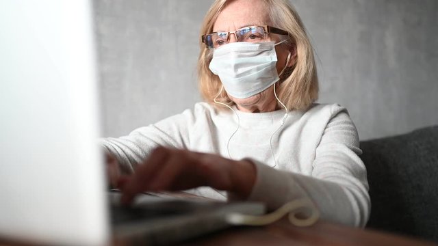 Technology, old age and people concept - lonely sad older senior woman in face medical mask working and making a video call with laptop computer at home during coronavirus COVID19 pandemic. Stay home