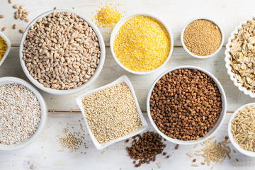 Selection of whole grains in white bowls - rice, oats, buckwheat, bulgur, porridge, barley, quinoa, amaranth, on white wood background