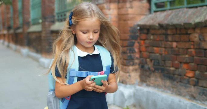 Adorable schoolgirl using modern smartphone while walking. Pretty kid with long blond hair and backpack going to school while looking and touching phone screen. Concept of igeneration.