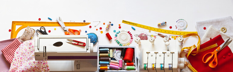 Sewing clothes. Panoramic top view on desktop: sewing machine, box with colored threads, overlock and accessories, scissors, buttons, needles, rulers and fabrics on white background. Banner