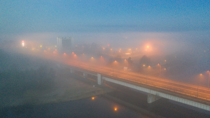 Obraz premium Aerial dawn colored view over heavy fog clad urban scenery of the Pärnu river and town with bridge, streetlamps and distant buildings