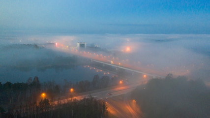 Aerial dawn colored view over heavy fog clad urban scenery of the Pärnu river and town with bridge, streetlamps and distant buildings