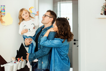 Mother, father and 3 years old girl spend time together at home. Beautiful young brunette mother hugging her husband and looking on blonde daughter. Family time during self isolation on quarantine.