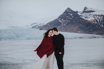 Wedding in Iceland. A guy and a girl in a white dress and a red jacket are walking on the ice against the backdrop of the mountains