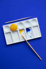 White palette with watercolor paints and brushes. Blue background. Instead of yellow, the egg yolk is used. Creative design and art. Top view and copy space