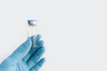 Doctors hand in blue rubber glove holding medicine bottle with blue cover and clear fluid on grey...