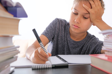 Schoolboy sitting among pile of books, textbooks, school exercise books on his desk and doing...