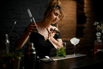 pretty girl bartender in black blouse carefully pours drink from jigger into mixing cup.
