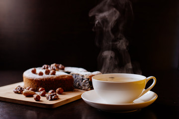 Biscuit with poppy seeds and nuts dusted with icing sugar. From a homemade cake cut a piece. Homemade sponge cake with poppy seeds and nuts, next to it is a cup with tea. Steam rises above the cup.