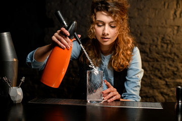 Young bartender girl attentively pours carbonated drink to glass using siphons.