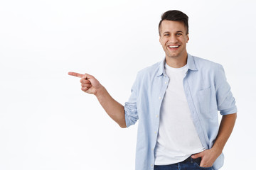 Portrait of happy, handsome masculine adult man pointing finger left and smiling camera, recommend service or product, best quality item, showing way to company banner, white background